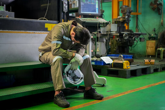 Factory worker industrial workshop fatigue stress safety helmet overtime manufacturing burnout, exhausted man sitting on step in machine shop, head down during break, workplace safety concept