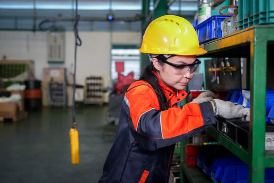 Factory worker woman hard hat safety goggle protective glove warehouse shelf inventory, focused technician sorting spare part in industrial storeroom for maintenance and logistics