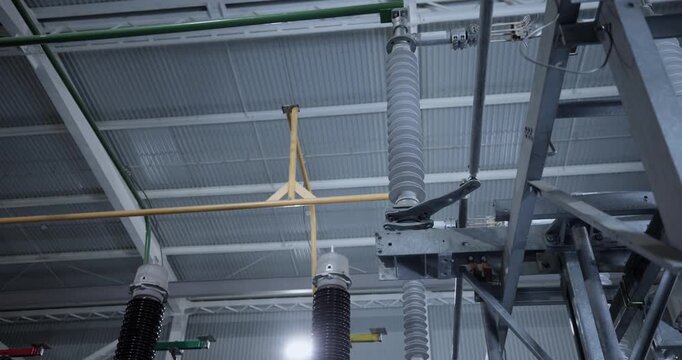 High voltage insulators and switchgear inside industrial substation under metal roof with overhead conduits.