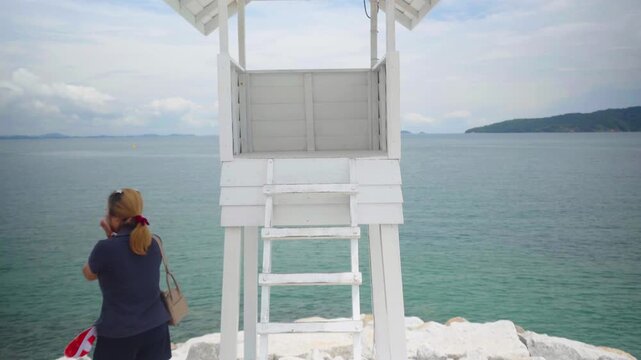 Asian female stands beside the iconic white wooden observation tower at Khao Laem Ya, gazing out over the beautiful turquoise waters of the Gulf of Thailand from the rocky white shoreline