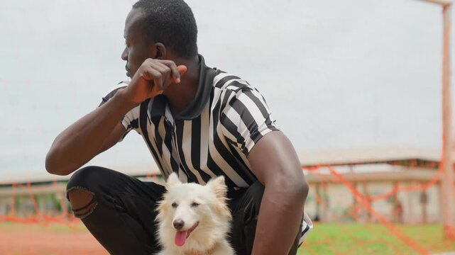 Black man crouching with dog in striped shirt near stadium fence, attentive trainer gesture, white fluffy puppy at knee, warm daylight, affectionate protective pose, urban sports field atmosphere,