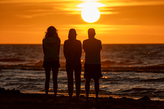 Saint-Come-De-Fresne, France - 08 10 2026: Two women and a man standing backlight in front of the sunset on the beach with the sea around at golden hour