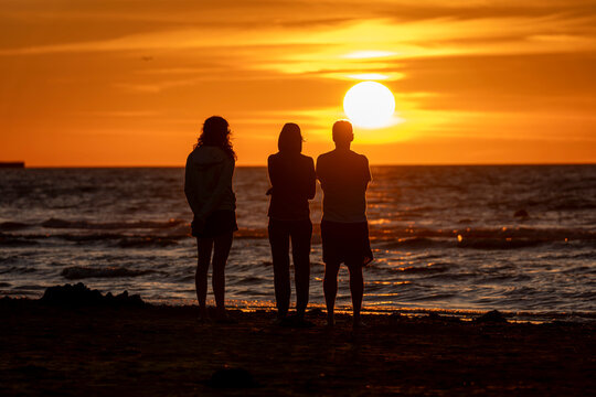 Saint-Come-De-Fresne, France - 08 10 2026: Two women and a man standing backlight in front of the sunset on the beach with the sea around at golden hour