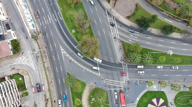 Top down aerial drone footage of a busy urban road intersection with moving vehicles, green areas and palm trees. Drone ascends vertically creating a smooth pull away shot.