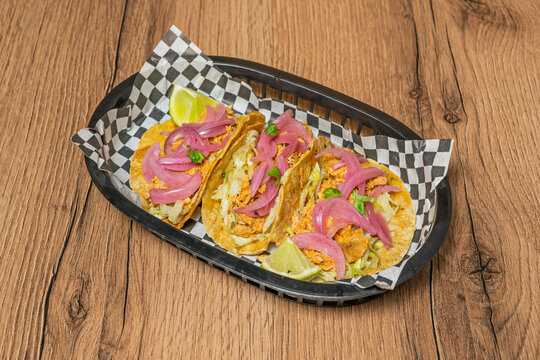 Cochinita pibil tacos topped with pickled red onion and lettuce, served with lime in a checkered basket.