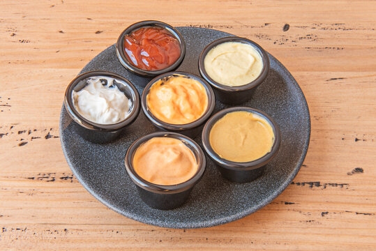 Six assorted dipping sauces in small black ramekins arranged on round dark plate, wooden table.