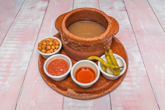 Cocido broth in a clay pot served with chickpeas, tomato sauce, oil, and pickled peppers in side bowls.