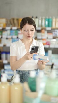 Thoughtful young female customer scanning QR code on box of ointment with phone to check price, read usage instructions and reviews before buying in local pharmacy . High quality 4k footage