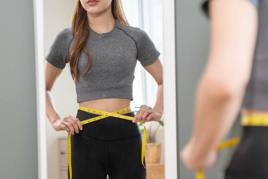 Weight lose, loss concept. Slim body asian young woman hand use tape measuring around waistline in fit sports. Girl looking reflect in mirror at home. Healthy nutrition, fitness for wellbeing beauty.