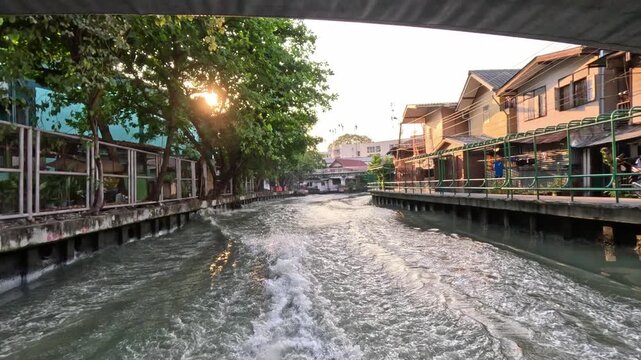 Wide shot of a river bay in Bangkok showing large waves and splashes from a fast-moving boat near low old residential houses by the canal.