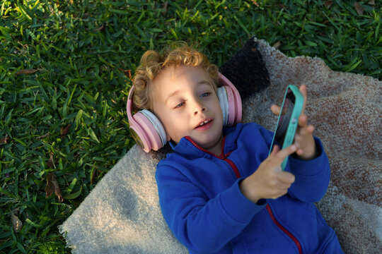 Toddler boy relaxing on grass, listening to music with headphones, exploring digital content on smartphone