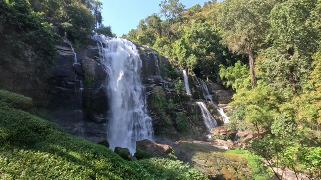 Wide low-angle shot of a large powerful waterfall and adjacent mini waterfalls surrounded by dense greenery, tall trees, and rocky cliffs in northern Thailand.