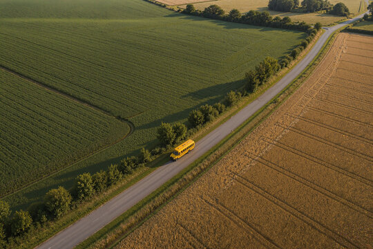 Aerial view of yellow school bus on rural road by green fields. School bus driving along scenic route amidst lush countryside and harvested fields.