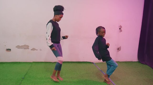 Black mother teaching child dance moves in bright studio on green turf upbeat coaching session with mother as instructor and child as eager student, barefoot steps, pink lighting and lively rhythm,