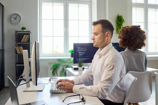 Developer programmer coding at computer in office. Two colleagues develop software as a team in a workplace with dual monitors, light and plants boosting focus. Teamwork and technology at work.