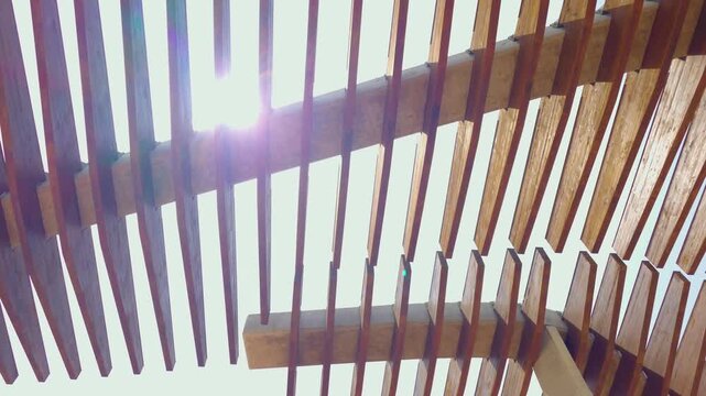 Sunlight streaming through wooden slatted canopy under bright summer sky. Sun rays between timber beams with lens flare. Modern pergola architecture, natural shade and outdoor geometric pattern