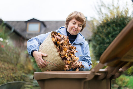 Senior woman emptying autumn leaf basket into compost bin outdoors