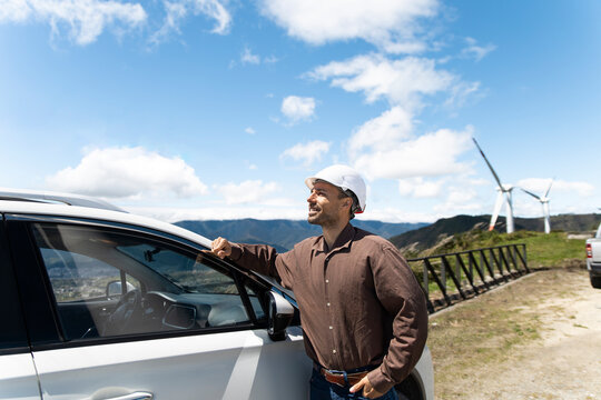 Male engineer leans on a car near a wind turbine in sunny Ecuador