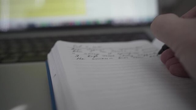 Man taking handwritten notes in notebook while working on laptop at home office desk. Concept of productivity, planning and focused remote work.