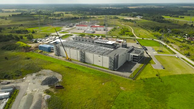 Drone footage showing the ongoing construction of a modern data center building, with a large crane installing rooftop cooling systems next to an electrical substation in a rural area