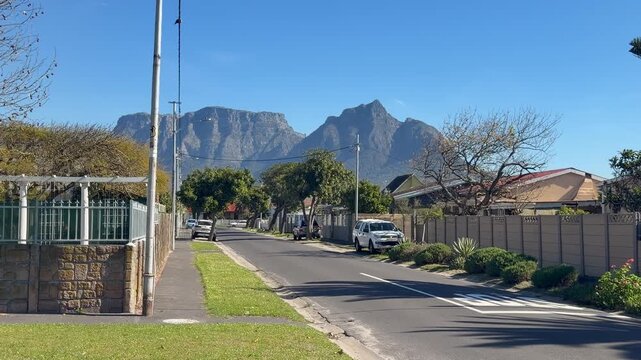 A tranquil residential street in Cape Town lined with walled properties, parked cars, and leafy trees, with Table Mountain and Devil&rsquo;s Peak rising dramatically in the background under clear blue skies