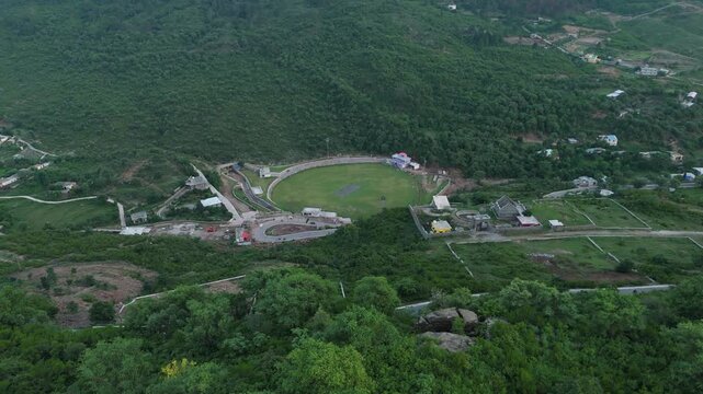 Birds eye view of a cricket ground surrounded by dense green pine trees and winding mountain roads as the camera pans down over the Margallah hills of South Asia