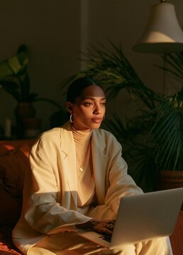Young woman sitting on a couch with a laptop in front of her. she is wearing a beige blazer and a gold necklace.