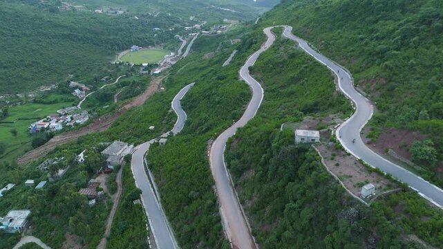 drone footage of vehicles and motorcycles navigating sharp hairpin turns on a steep winding serpentine mountain road overlooking a green valley and cricket stadium in the Margallah Hills of Pakistan