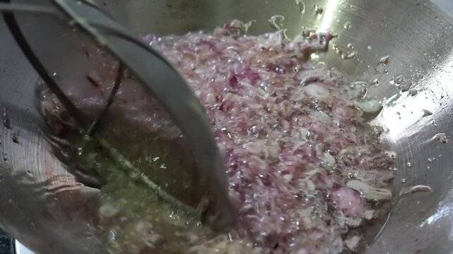 Medium close up video of a cook using a mesh strainer utensil to stir shallots frying in hot oil within a wok in slow motion.
