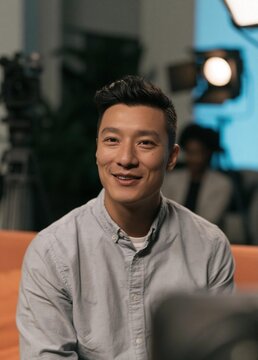 Portrait of a young asian man sitting on an orange couch. he is wearing a light grey button-down shirt and has short dark hair. he has a big smile on his face and is looking directly at the camera.