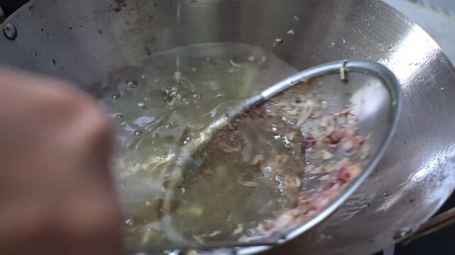 Slow motion footage showing sliced red shallots being lowered into hot oil using a metal strainer mesh and stirred slowly in a large wok during food preparation.
