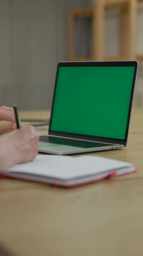 Woman Working On Laptop Computer with Green Mock Up Screen At Home Office Space. Female Freelancer Browsing Green Laptop Screen Writing Information in Notebook. Vertical Video