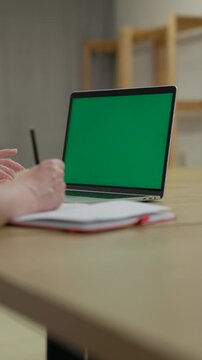 Woman Working On Laptop Computer with Green Mock Up Screen At Home Office Space. Female Freelancer Browsing Green Laptop Screen Writing Information in Notebook. Vertical Video