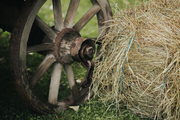 Wooden wagon wheel positioned beside a round bale of hay on green grass, showcasing rustic farm elements in a natural outdoor setting © Emvats