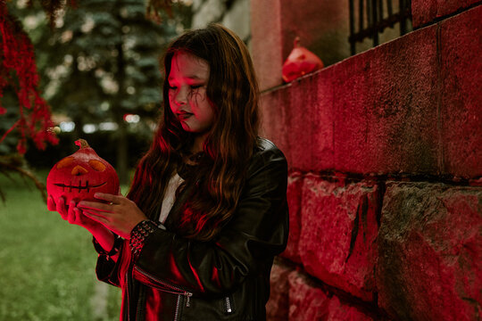 Asian teenager girl wearing spooky makeup holding carved jack o lantern pumpkin outdoors during Halloween celebration. Image suitable for seasonal marketing campaigns