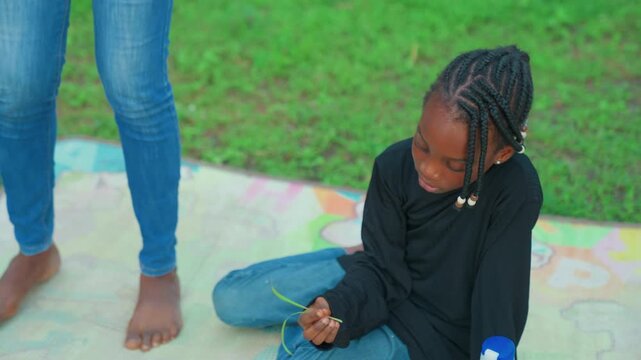 Mother and child on blanket outdoors young girl with braided hair sits crosslegged on patterned blanket while barefoot mother stands nearby child examines small object with focused curiosity, denim