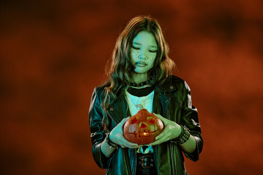 Asian teenager girl wearing spooky makeup holding carved jack o lantern pumpkin during halloween celebration. Image suitable for autumn holiday marketing campaigns, medium shot