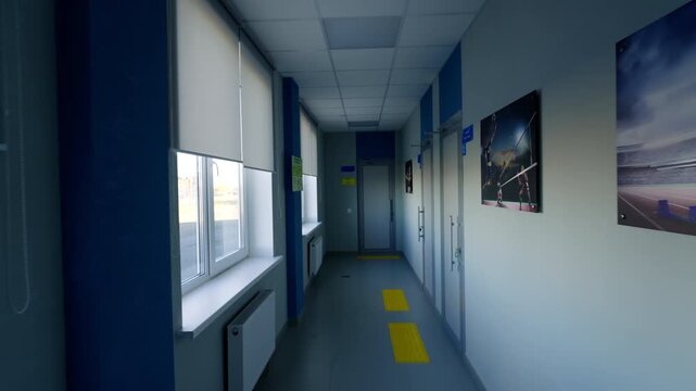 Narrow corridor features windows with blinds opposite posters of running athletes. Empty hallway looks quiet and well-lit during daytime