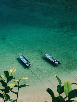 High angle top view of two blue traditional wooden fishing boats floating on crystal clear turquoise ocean water with green leaves frame