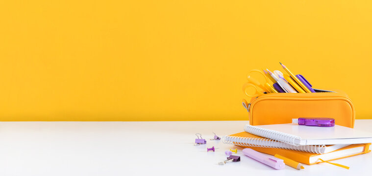 Yellow Pencil Case and Notebooks on a White Desk Surface Against a Yellow Wall. Minimalist Office Background with Scissors, School Supplies, and Large Copy Space for Text. Panoramic Banner Layout.