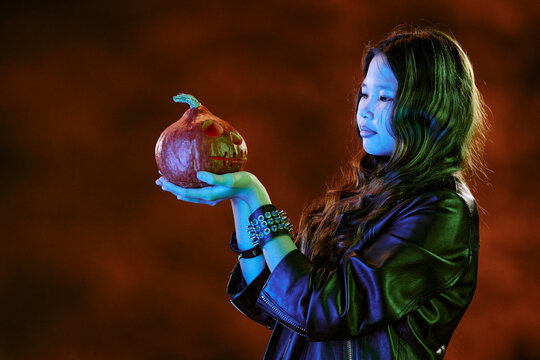 Asian teenager girl holding carved Halloween pumpkin, celebrating autumn holiday. Image suitable for seasonal marketing campaigns, festive event promotions and youth lifestyle advertisements