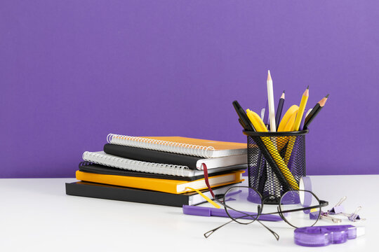 Stack of Notebooks and Yellow Stationery in a Mesh Holder on a White Desk Against a Purple Wall. Minimalist Office Background with Eyeglasses, School Supplies, and Copy Space. Horizontal Banner.