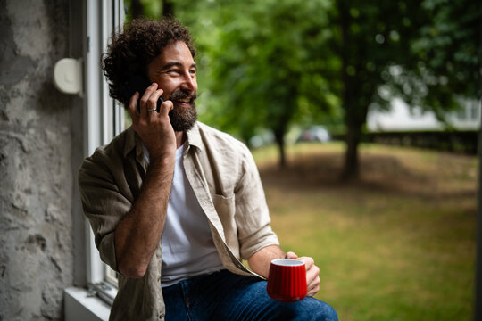 Happy man having phone call holding mug by window