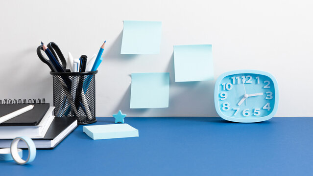 Blue Alarm Clock and Sticky Notes on a White Wall Above a Blue Desk. Minimalist Office Workplace Background with Notebooks, Stationary Holder, Pencils, and Copy Space for Text. Banner Layout.