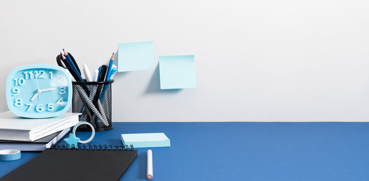 Blue Alarm Clock and Sticky Notes on a White Wall Above a Blue Desk. Minimalist Office Workplace Background with Notebook, Stationery Holder, Pencils, and Large Copy Space for Text. Banner Layout.
