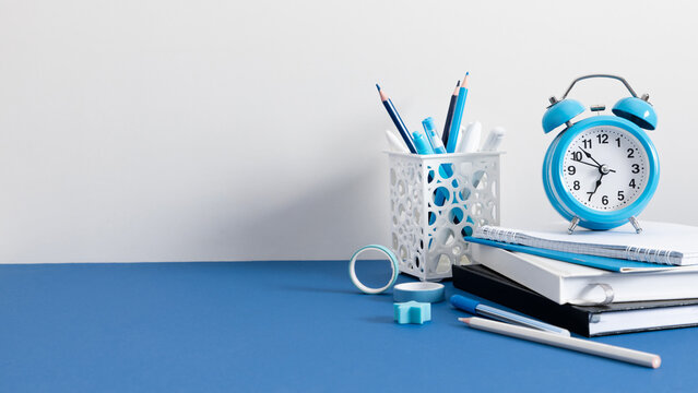 Blue Alarm Clock and School Stationery on a Blue Desk Against a White Wall. Minimalist Office Workplace Background with Notebooks, Stack of Books, and Colored Pencils in a White Holder. Banner.