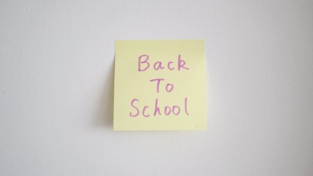A person's hand pressing a square yellow sticky note onto a plain white wall. The note features the words "Back To School" written in purple handwritten script.