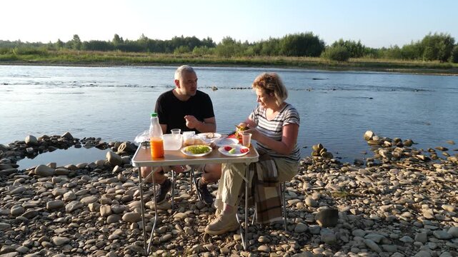 Couple spends leisure time by the river, tasting food and drinks during a peaceful summer outing in nature.
