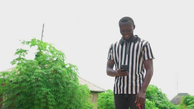 Black man scrolling phone by bushes. Urban content creator and everyday communicator standing beside dense green foliage, thumbs navigating smartphone feed, casual striped shirt, focused expression,