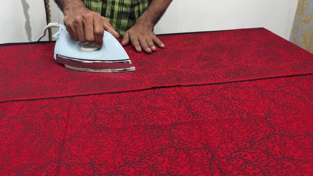 Close-up of a tailor or laundry worker meticulously ironing red traditional batik fabric or table cloth using an electric steam iron on a work table.
